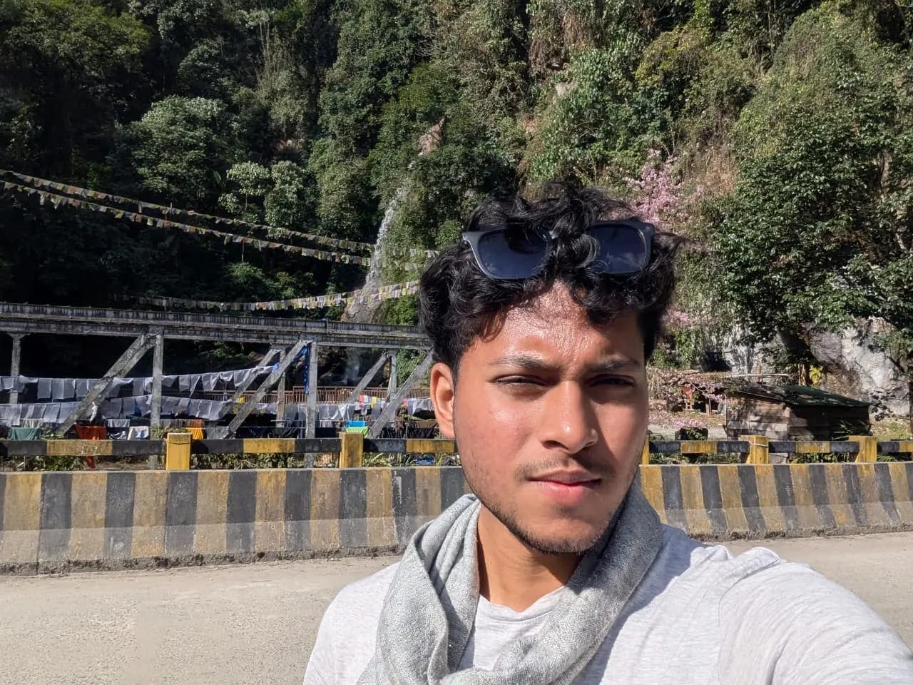 Aman at a bridge strung with prayer flags, a waterfall behind